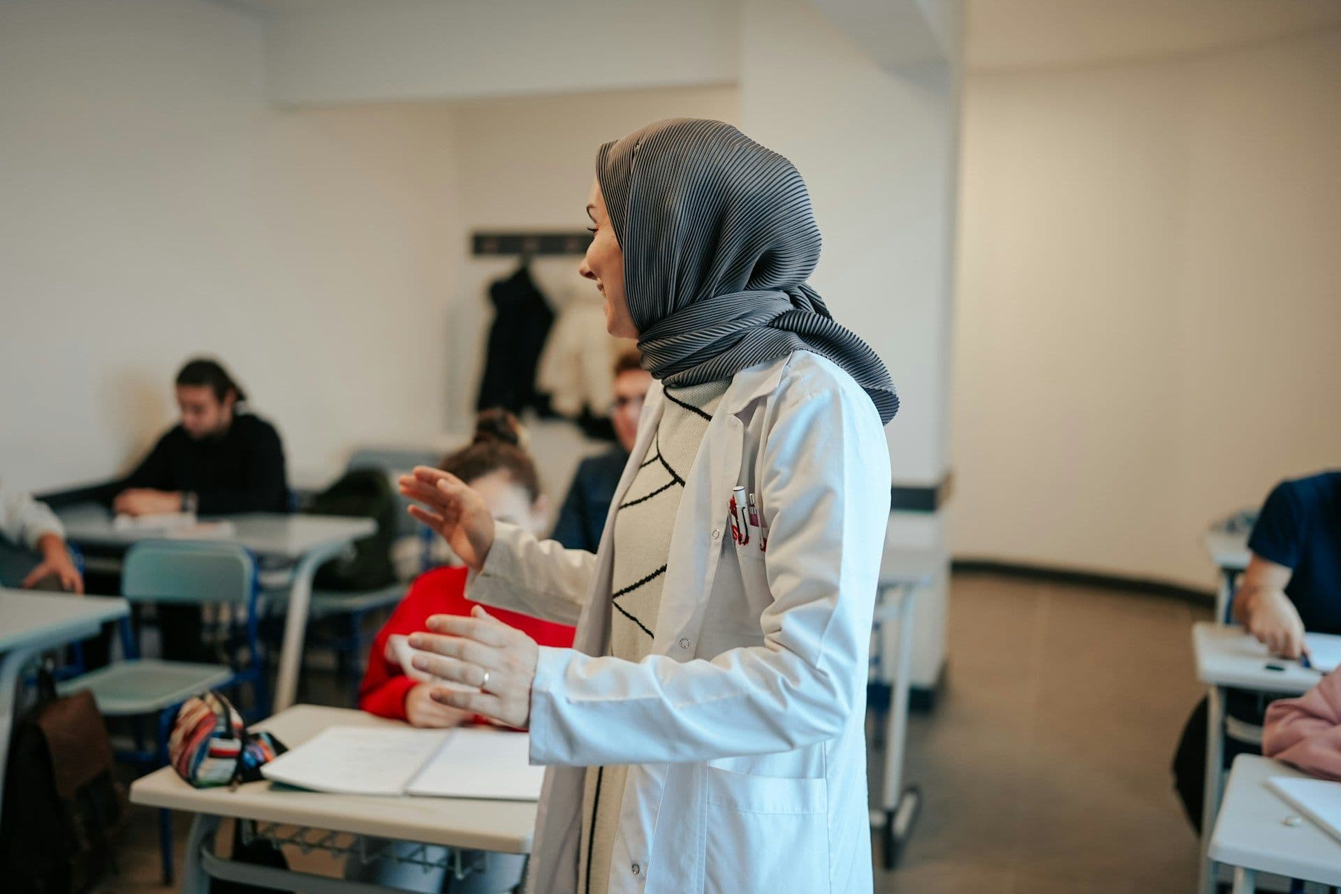 A teacher in a headscarf talking with students in a bright secondary classroom